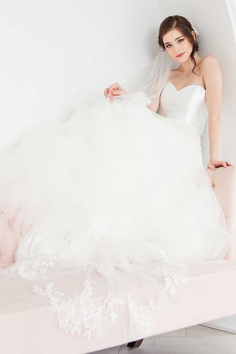 Bride seated wearing hand embroidered floral Indra Cathedral Veil with train cascading over the side of the sofa.