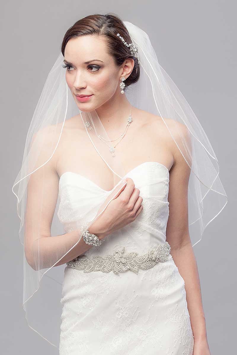 Modern minimalist bride looking to the side holding the edge of classic two tier fingertip wedding veil with blusher. Opulence Two Tier Fingertip Veil by Laura Jayne Accessories Toronto. Handcrafted in Canada.