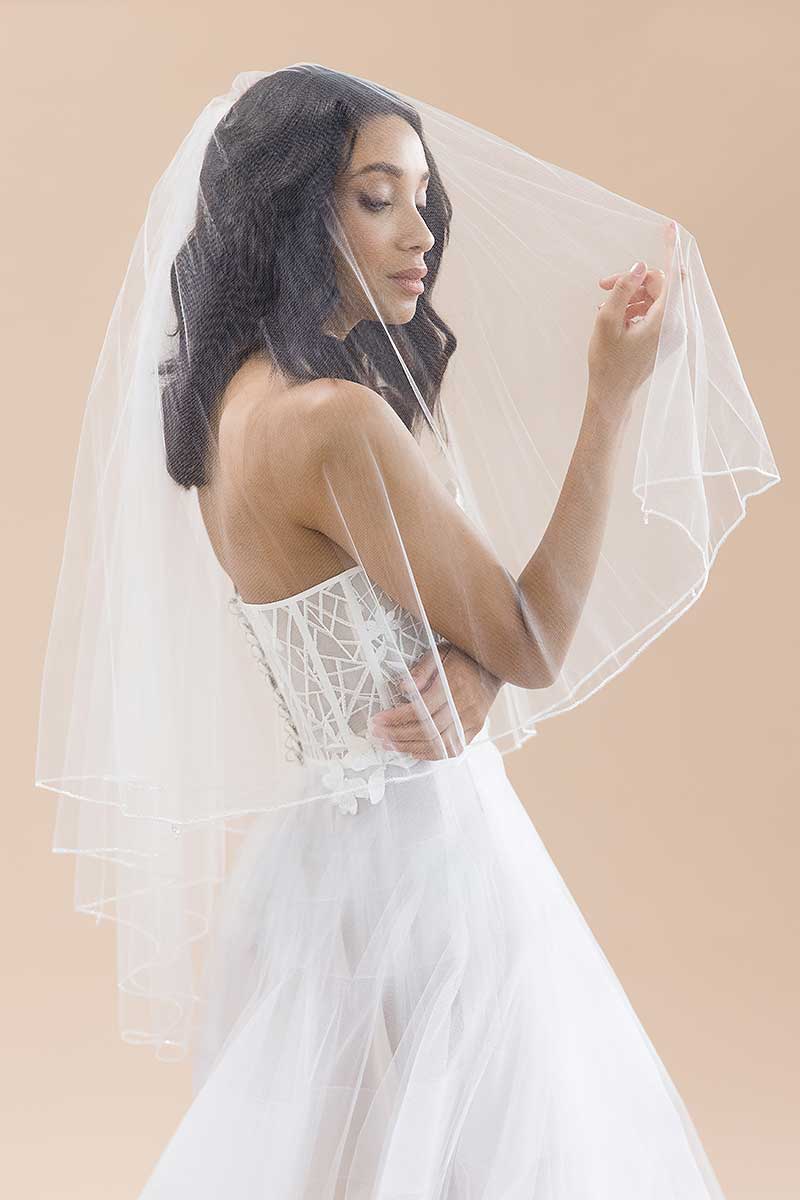 Bride wearing strapless wedding gown and sheer blusher veil with pearl and crystal details along the border. Jess Beaded Edge Fingertip Veil by Laura Jayne Accessories. Handcrafted in Toronto, Canada.
